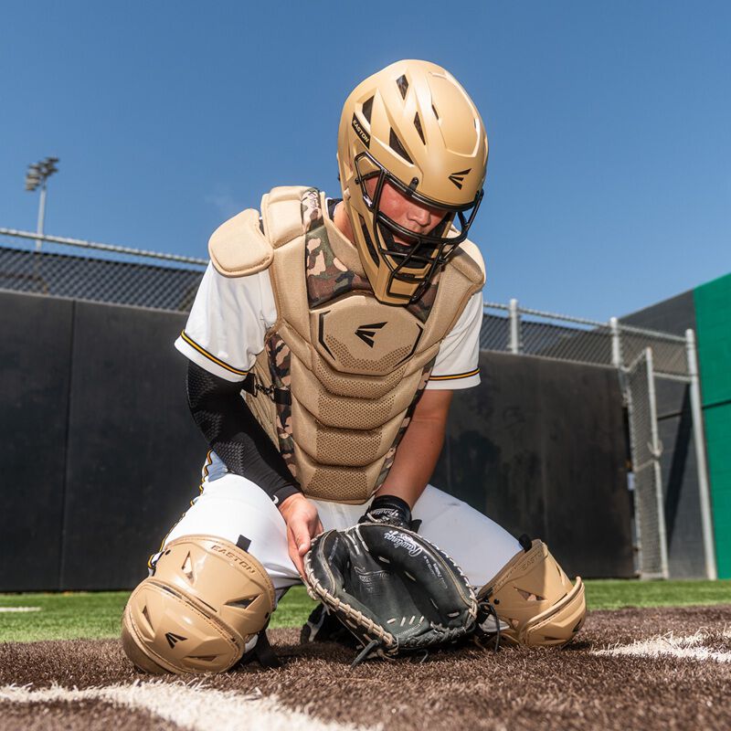 Youth catcher wearing tan and camo Easton Mavrix catcher's gear kneeling behind home plate with mitt down